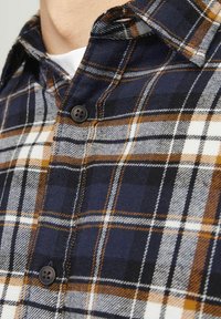 Košulja karo uzorka u mornarici i narančastoj boji, izrađena od flanelskog materijala. Ima dugmad za zatvaranje s tamnim dugmadima i klasičan dizajn ovratnika.