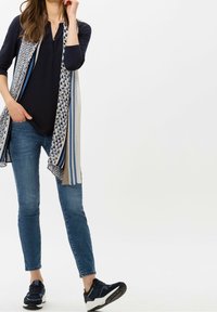 Mehrfarbiger, gemusterter Schal über einem schwarzen Oberteil. Die Jeans sind hellblau, und die Schuhe sind dunkelblau mit weißen Akzenten und strukturierten Sohlen.