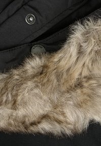 Zwarte stof met een faux fur rand in tan. Beschikt over een glanzende drukknoop en een textuur oppervlak, wat contrast biedt in materiaal en kleur.