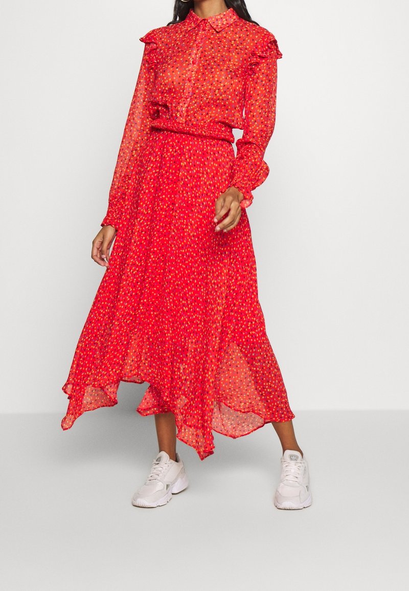 Femme portant une robe longue à manches longues rouge fleurie avec col et ourlet à volants, associée à des baskets blanches sur un fond uni.