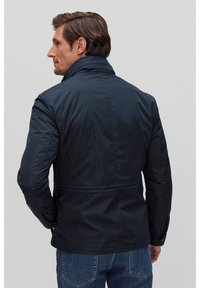 Tummansininen kevyt takki, jossa on pystykaulus, virtaviivainen muotoilu, hienovaraisesti teksturoitu kangas ja kapea leikkaus vyötäröllä.