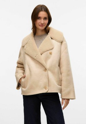 Kvinde iført en beige shearlingjakke over en lys brun sweater og mørke bukser, stående med den ene hånd i lommen.