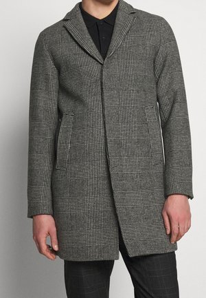 Mann trägt einen grauen karierten Mantel mit Klappentaschen über einem schwarzen Hemd mit Kragen und dunkel karierten Hosen vor einem einfarbigen Hintergrund.