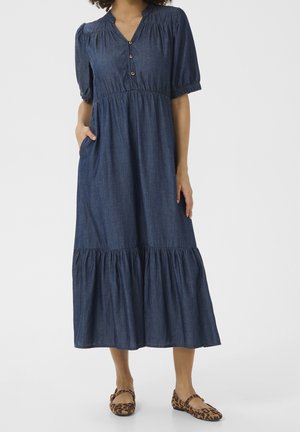 Kvinde iført en blå denim midi-kjole med pufærmer, knappet V-hals, sidelomme og leopardprint flade sko, stående på hvid baggrund.