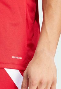 Rotes Sportshirt aus glattem Stoff mit einem Etikett, auf dem "AEROREADY" steht. Mit einem kontrastierenden weißen Akzent am Saum.