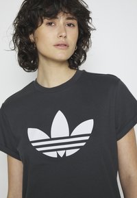 Juodas medvilnės marškinėliai su dideliu baltu trefoil logo ir trimis horizontaliomis juostelėmis. Apvalus iškirpimas ir trumpomis rankovėmis.