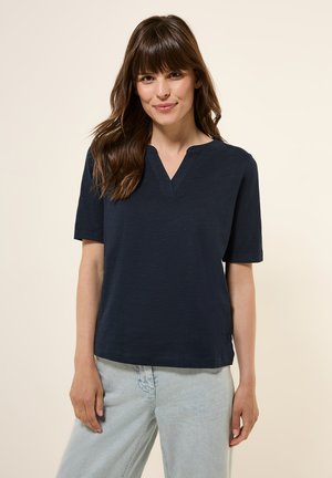 Dark navy short-sleeve top with a V-neckline, made from a textured fabric. Features a relaxed fit and subtle side slits. Worn with light jeans.