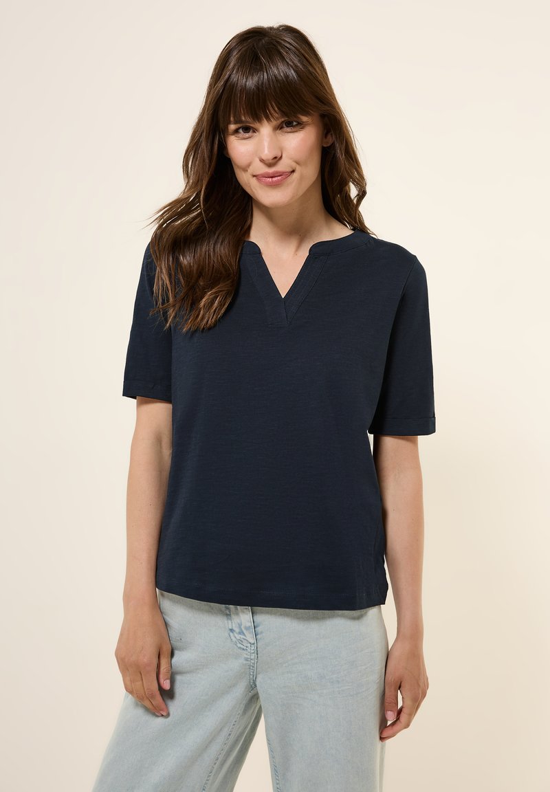 Haut court à manches courtes bleu marine foncé avec un décolleté en V, fabriqué à partir d'un tissu texturé. Présente une coupe décontractée et de subtils fentes latérales. Porté avec un jean clair.