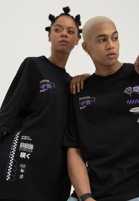 To modeller som har på seg svarte grafiske t-skjorter med hvit og lilla tekst og symboler, poserer foran en ensfarget hvit bakgrunn.