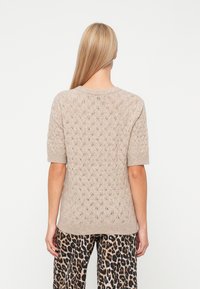 Γυναίκα με μακριά ξανθά μαλλιά, φορώντας μπεζ κοντομάνικη πλεκτή μπλούζα με σχέδιο φύλλων και παντελόνι λεοπάρ. Στηρίζει τον εαυτό της σε λευκό φόντο.