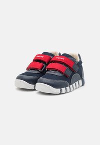 Scarpe per bambini blu in materiale sintetico con fascette in Velcro rosse, suola bianca e design testurizzato. Caratterizzate da una costruzione flessibile e traspirante.