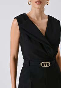 Tutina nera con scollo a V profondo, spalle strutturate e vita con cintura. L'accessorio in metallo dorato sulla cintura aggiunge contrasto.