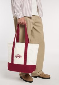 Osoba ubrana w khaki spodnie i beżowe buty trzyma kremową i bordową torbę Dickies za uchwyty na jednolitym tle.