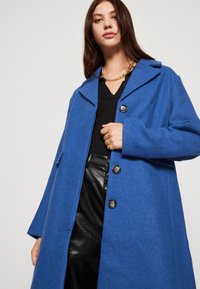 Koboltblå ullfrakk med snippelapper, fire svarte knapper og sidelommer. Modellen har på seg en svart topp og skinnbukser.