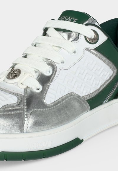 Gros plan d'une sneaker Versace avec des lacets blancs, des panneaux verts et argentés, un motif clé grec en relief et un emblème métallique Méduse près de la pointe.