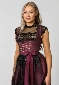 Bordeauxfarbener Dirndl mit Spitzeakzenten, durchscheinendem schwarzen Mieder, tailliertem Schnitt, floralen Details und Satinschleifen-Gürtel. Textil mit strukturiertem Stoff und filigranen Mustern.