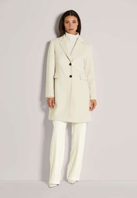 Vrouw in een crèmekleurige lange jas met zwarte knopen, witte coltrui, witte broek en witte puntige schoenen, staand tegen een beige muur.