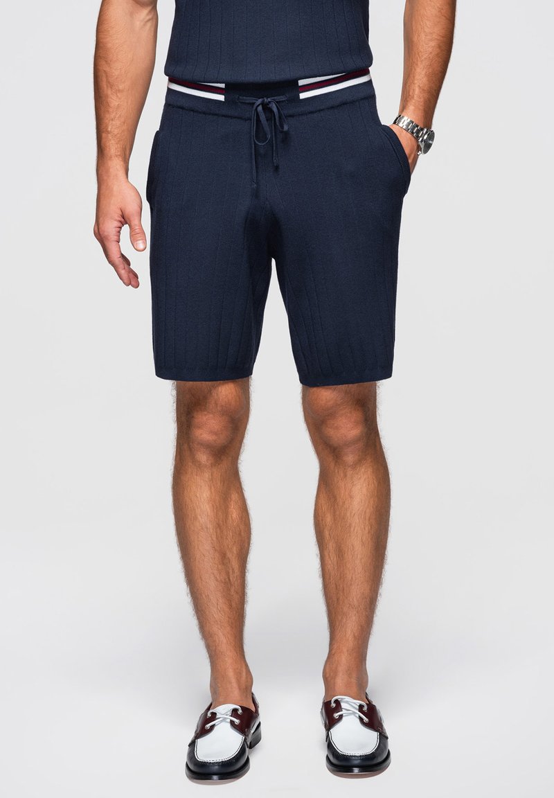 Homme portant un short en maille bleu marine avec cordon de serrage et poches, associé à des chaussures bateau bicolores blanc et bordeaux, debout avec une main dans la poche.
