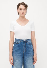 Junge Person mit kurzen, gewellten Haaren, die Hängeohrringe, ein weißes V-Ausschnitt-T-Shirt und hochgeschnittene blaue Jeans trägt und vor einem weißen Hintergrund steht.