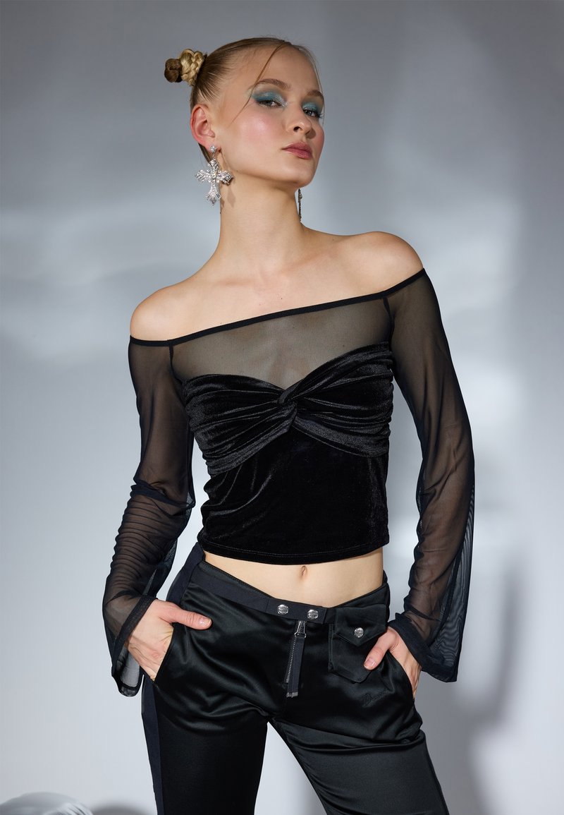 Crni baršunasti crop top s prozirnim dugim rukavima, s prekrivenim prednjim detaljem. Model nosi crne hlače sa zatvaračem i dekorativnim gumbima.