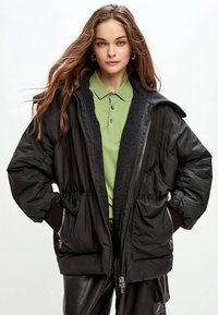 Tânără cu păr lung poartă o jachetă neagră matlasată peste o cămașă verde cu guler și pantaloni negri, stând pe un fundal alb simplu.