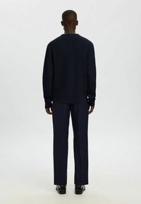 Uomo di spalle che indossa un maglione texturato blu scuro, pantaloni abbinati e scarpe nere lucide su uno sfondo chiaro e semplice.