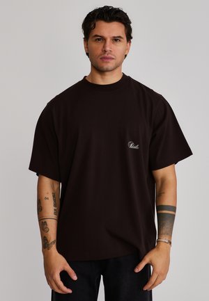 Mand med mørkt hår iført en sort oversized T-shirt med lille hvidt logo, der viser tatoveringer på begge underarme, stående foran en ensfarvet baggrund.