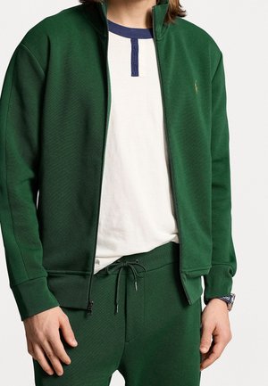 Green zip-up jacket made of textured fabric, paired with a white shirt featuring a blue neck accent. Slim fit with drawstring pants.