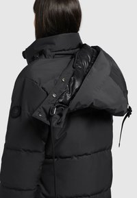 Person with long dark hair wearing a black padded jacket with an adjustable hood partially unzipped at the back.