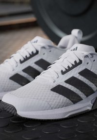 Ζευγάρι λευκά και μαύρα αθλητικά παπούτσια Adidas τοποθετημένα σε μαύρο λαστιχένιο δάπεδο γυμναστηρίου με εξοπλισμό άρσης βαρών στο φόντο.