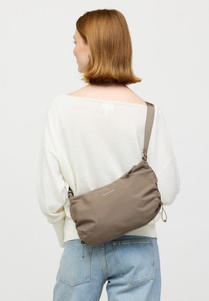 Frau mit kurzem roten Haar, die einen weißen Pullover und hellblaue Jeans trägt, hat eine taupefarbene Umhängetasche auf dem Rücken.