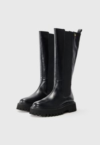 Bottes en cuir noires, hautes, avec des semelles épaisses à crampons et des panneaux élastiques sur les côtés, sur un fond blanc uni.