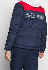 Marineblauer Steppjacke mit rotem Schulterpaneel und weißem Logo. Verfügt über eine gesteppten Textur und elastische Bündchen für Wärme und Komfort.