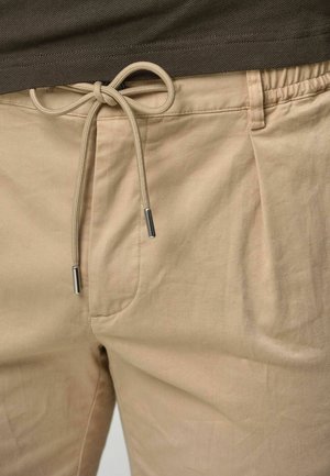 Pantalones beige con cintura elástica, cordón ajustable y puntas metálicas plateadas. La tela parece suave, con una textura lisa y pocas arrugas.