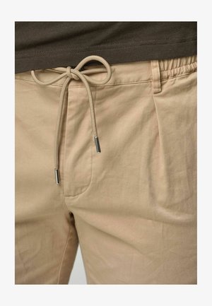 Pantaloni beige con vita elastica, coulisse regolabile e punte in metallo argento. Il tessuto appare morbido, con una texture liscia e pieghe minime.