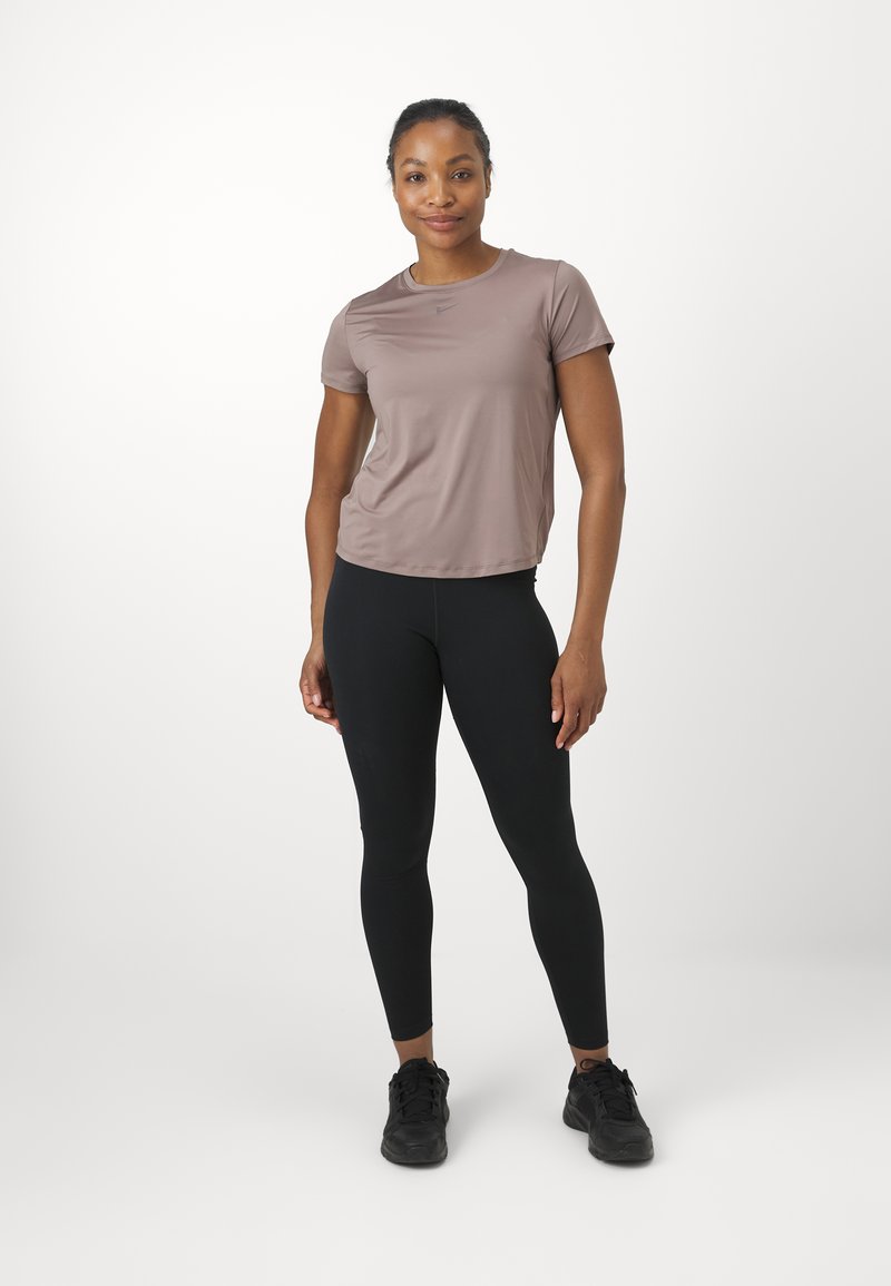 Maglietta atletica corta di colore marrone chiaro abbinata a leggings neri. La maglietta presenta un collo rotondo e una texture liscia. Calzature sono scarpe da ginnastica nere.