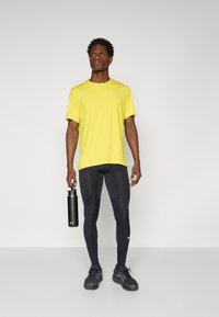 T-shirt de sport à manches courtes jaune, leggings noirs ajustés, chaussures de sport noires, tenant une bouteille d'eau noire. Design simple et fonctionnel.