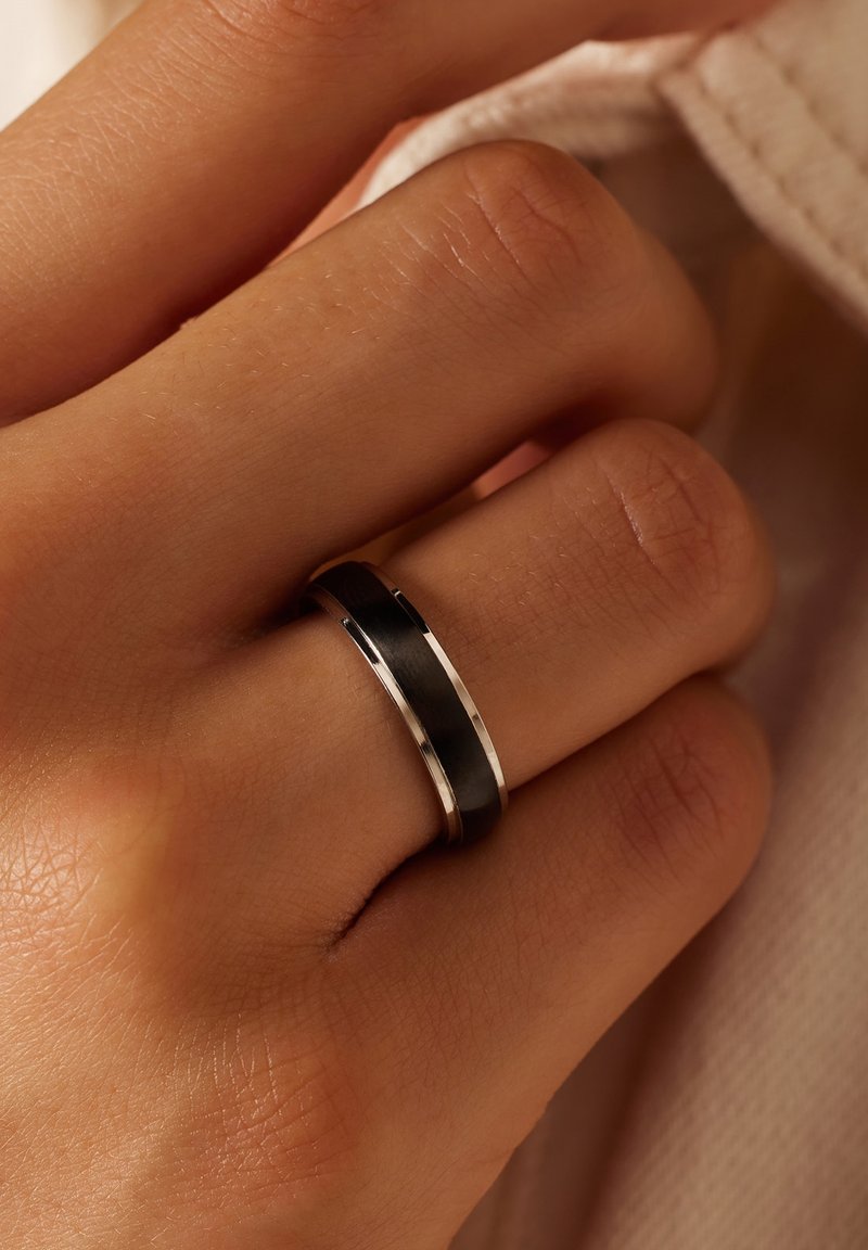 Zwarte en zilveren ring op een hand; heeft een gladde zwarte band met een zilveren rand. De setting is gepolijst, met een slank ontwerp.