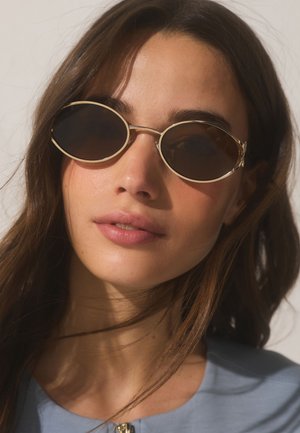 Mladá žena s dlhými hnedými vlasmi, ktorá má na sebe oválne tmavé slnečné okuliare a svetlomodrý top na jednofarebnom pozadí.