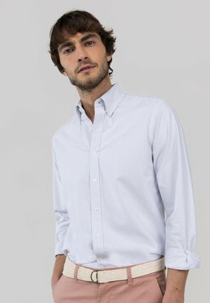 Hombre con barba clara que lleva una camisa de rayas azul claro abotonada y pantalones rosa claro con cinturón blanco tejido posando frente a un fondo liso.