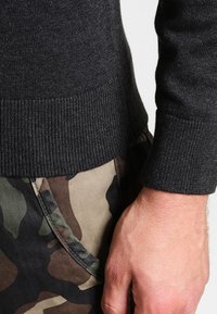 Close-up of a hand by a hip wearing a dark gray knitted sweater and camouflage-patterned pants.