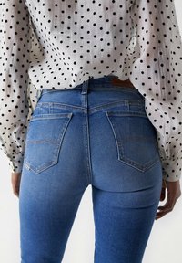 Jean bleu taille haute en denim avec une texture lisse, dotée de deux poches arrière et de coutures contrastées. Blouse à pois blancs visible.