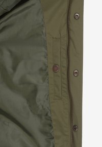 Innenseite einer olivgrünen Jacke mit glatter Textur, ausgestattet mit einer seitlichen Tasche mit Klappe, metallischen Knöpfen und einem Reißverschluss.