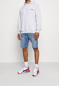 Sudadera gris con capucha y logo, pantalones cortos de mezclilla azul, y zapatillas blancas con detalles en rojo y azul, llevadas con calcetines blancos de caña corta.