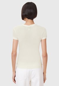 T-shirt a maniche corte di colore crema con un design aderente, collo tondo e texture liscia. Presenta un piccolo logo sulla parte alta della schiena.