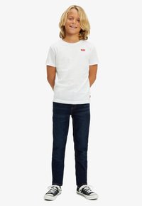 Biała bawełniana koszulka z czerwoną logo Levi's na piersi, w połączeniu z ciemnoniebieskimi dżinsami i czarnymi trampkami z białymi sznurowadłami. Dziecko stoi z rękami za plecami.