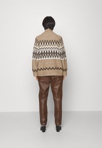 Maglione lavorato a maglia color beige con motivi geometrici bianchi e marroni; indossato con pantaloni di pelle marrone. Presenta un colletto alto e una vestibilità oversized.
