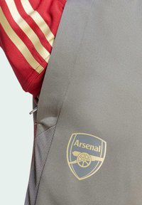 Jersey atlético gris con mangas rayadas en rojo y dorado, con un logo dorado de Arsenal en el lado derecho. Textura suave, diseño deportivo.