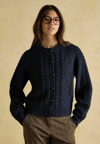 Tums zilas adīta kardigāna ar kabeles rakstiem, ar apaļu kakla izgriezumu un pogu aizdari, apvienots ar raibu biksēm.