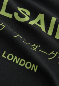 Melnas auduma t-krekls ar drosmīgiem zaļiem burtiem un simboliem. Teksts ietver vārdu "LONDON" un dažādas valodu rakstības. Tekstūrēta virsma.